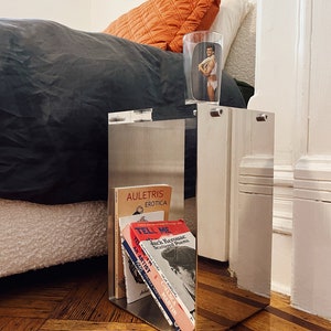 May include: A silver mirrored side table with a stack of books on the shelf. The books are titled "Auletris Erotica", "Tell Me", "Jack Kerouac Scattered Poems", "An Artist's Life", and "Chelsea Martin".