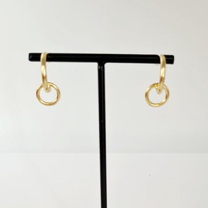 Op de afbeelding: Een paar gouden creolen met een kleinere cirkel die aan de creool is bevestigd. De oorbellen worden gepresenteerd op een zwarte standaard met een decoratieve basis.