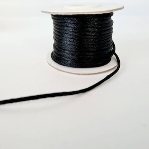Può includere: Un rocchetto di cordoncino cerato nero, avvolto attorno a un rocchetto di cartone bianco. Il cordoncino è sottile e ha una finitura liscia e lucida.