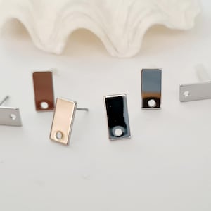 Puede incluir: Cuatro pares de bases rectangulares para pendientes con un agujero en el centro. Las bases son plateadas, doradas, negras y blancas.