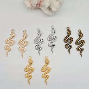 Könnte beinhalten: Ein Set mit sechs Schlangen-Anhängern in Gold-, Silber- und Bronze-Finish. Die Schlangen-Anhänger sind mit strukturierten Schuppen verziert und perfekt, um Schmuckprojekten einen Hauch von Verspieltheit zu verleihen.