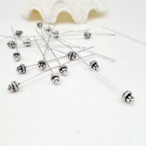 Peut inclure: Un tas d'épingles à tête argentées avec des sommets décoratifs. Les épingles sont éparpillées sur une surface blanche.
