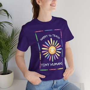以下が含まれることがあります： 紫色のTシャツ。笑顔の太陽のデザインと「HAPPY to TEACH TINY HUMANS」の文字。太陽は笑顔で、カラフルなクレヨンのフレームで囲まれています。