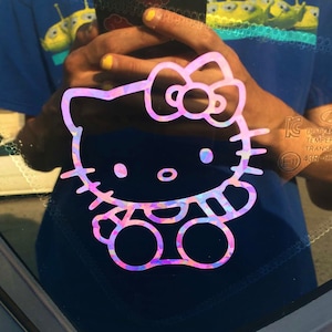 May include: A holographic Hello Kitty sticker, featuring a pink, purple, and blue color scheme. The sticker is applied to a window, with the iconic character's face, bow, and body clearly visible. The sticker is a fun and decorative item.