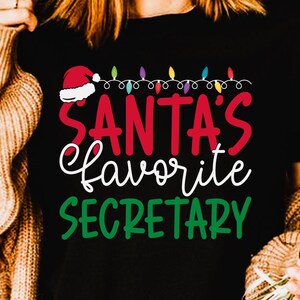 Może przedstawiać: Czarny t-shirt z napisem "SANTA'S favorite SECRETARY" w kolorach czerwonym, białym i zielonym. Projekt zawiera czapkę Świętego Mikołaja i łańcuch światełek. Produkt cyfrowy do natychmiastowego pobrania.