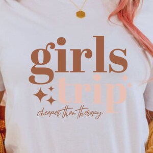 May include: White t-shirt with brown text that says "girls trip" and smaller pink text that says "cheaper than therapy".