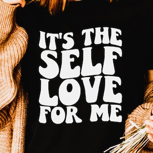 May include: A black t-shirt with white text that reads "It's the Self Love for Me" in a retro font style. The shirt is being worn by a person with brown hair and a brown cardigan.