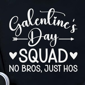 Puede incluir: Camiseta negra con el texto blanco "Galentine's Day SQUAD NO BROS, JUST HOS". La camiseta tiene las mangas dobladas y un nudo en el bajo. También se ven unas zapatillas deportivas blancas y negras y una pequeña planta.