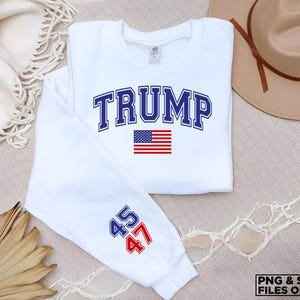 以下が含まれることがあります： 白いスウェットシャツに、青い文字で「TRUMP」と書いてあり、その下に小さなアメリカ国旗があります。袖には青い文字で「45」と「47」が書かれています。