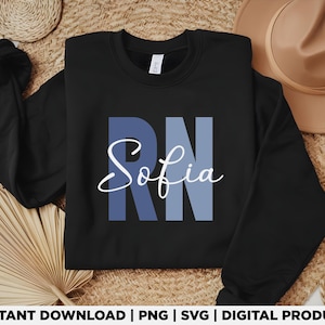 Pode incluir: Camisola preta de gola redonda com as letras "RN" em azul e o nome "Sofia" em escrita branca. A camisola é exibida em uma superfície neutra com uma esteira de palha tecida e um chapéu bege.
