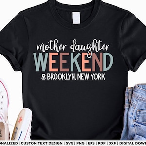May include: Black t-shirt with the text "mother daughter WEEKEND" in white, teal, and peach. Below the text is "BROOKLYN, NEW YORK" and a location pin icon. White Converse sneakers and denim jeans are visible.