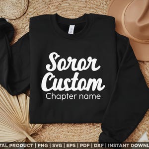 Puede incluir: Una sudadera negra con las palabras "Soror Custom" en escritura blanca. Debajo, el texto "Chapter name" también está en blanco. La sudadera se muestra sobre un fondo neutro con un sombrero y elementos tejidos.
