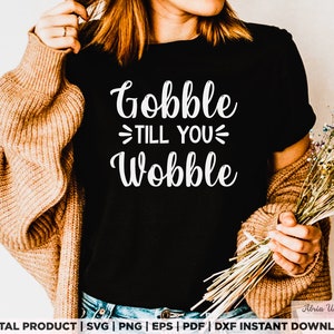 May include: Black t-shirt with the white text "Gobble Till You Wobble". The shirt is part of a digital product collection. The image also includes a brown cardigan and a bouquet of dried flowers.