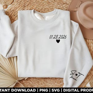 May include: A light gray sweatshirt with the date "01.28.2026" and a black heart printed on the chest. The sleeve features the text "S & T forever" in black script. The sweatshirt is displayed on a woven surface.