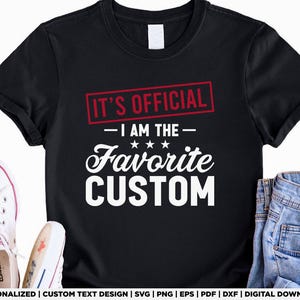 May include: Black t-shirt with the text "IT'S OFFICIAL - I AM THE - Favorite CUSTOM" in white and red. The shirt is paired with white sneakers and blue jeans. The design is for personalized custom text.