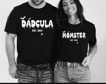May include: Black t-shirts with white text. One shirt says "Dadcula" with bats, a spider, and "Est. 2024." The other says "Momster" with bats, a spider, and "Est. 2024." Both shirts have a dripping effect on the text.