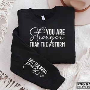 May include: Black sweatshirt with white text that reads "You are stronger than the storm" and "This too shall pass" on the sleeve.