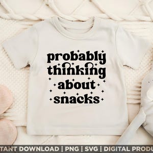 Pode incluir: Uma camiseta creme com a frase "probably thinking about snacks" em preto, com uma fonte estilizada. Pequenas estrelas pretas circundam o texto. A camiseta é exibida em uma superfície macia e texturizada.