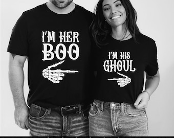 May include: Two black t-shirts with white text and skeleton hand graphics. One says "I'M HER BOO" with an arrow, the other says "I'M HIS GHOUL" with an arrow. The shirts are worn by two people, a man and a woman.