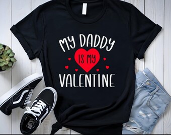 May include: Black t-shirt with white text that reads "My Daddy is My Valentine" with a red heart in the middle. There are small red hearts around the heart.