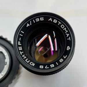 May include: A black camera lens with the text "4/135 AUTOMAT" and "KOMINEF-II" printed on the side. The lens has a black metal body and a clear glass lens.