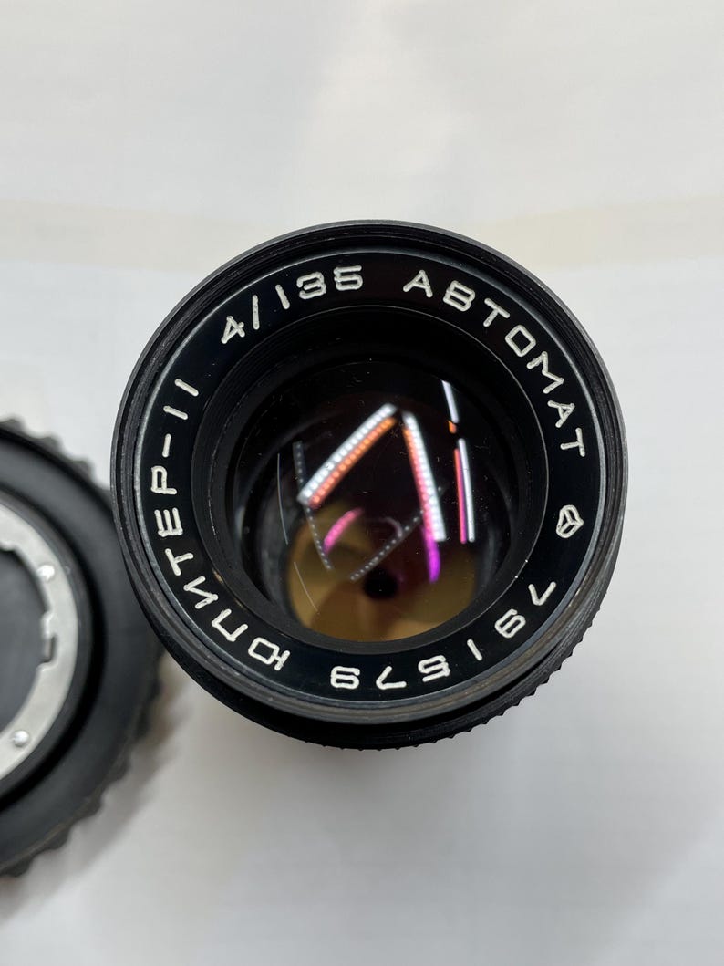 May include: A black camera lens with the text "4/135 AUTOMAT" and "KOMINEF-II" printed on the side. The lens has a black metal body and a clear glass lens.