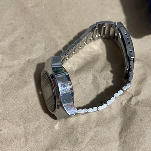 以下が含まれることがあります： 長方形の金属製ケースとマッチするリンクブレスレットを備えたシルバーカラーのデジタル腕時計。文字盤はダークカラーでデジタル表示となっており、金属製のバンドには手首にしっかりと固定するための折りたたみ式クラスプ機構が付いています。