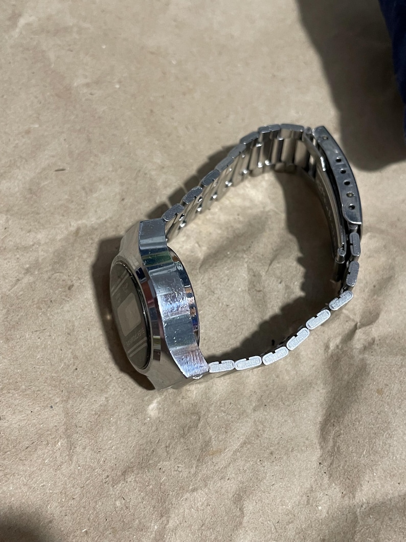 以下が含まれることがあります： 長方形の金属製ケースとマッチするリンクブレスレットを備えたシルバーカラーのデジタル腕時計。文字盤はダークカラーでデジタル表示となっており、金属製のバンドには手首にしっかりと固定するための折りたたみ式クラスプ機構が付いています。