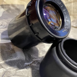 Op de afbeelding: Een zwarte cameralens met de tekst "TPVЛИET 1:3 f=10,5mm" is gedeeltelijk zichtbaar. De lens heeft een reflecterend oppervlak met paarse en gele tinten. Een zwarte lensdop ligt in de buurt.
