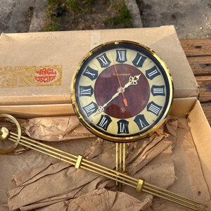 May include: A vintage clock with a gold-colored frame and black Roman numerals on a burgundy face. The clock includes a gold-colored pendulum and is displayed in its original cardboard box. The box has text in a foreign language.