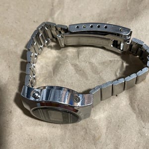 以下が含まれることがあります： 長方形の金属製ケースとマッチするリンクブレスレットを備えたシルバーカラーのデジタル腕時計。時計のケース側面には2つのプッシュボタンがあり、調整可能な金属製バンドには折りたたみ式の留め金機構が付いています。