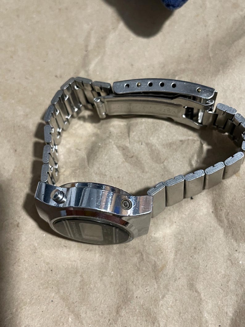以下が含まれることがあります： 長方形の金属製ケースとマッチするリンクブレスレットを備えたシルバーカラーのデジタル腕時計。時計のケース側面には2つのプッシュボタンがあり、調整可能な金属製バンドには折りたたみ式の留め金機構が付いています。
