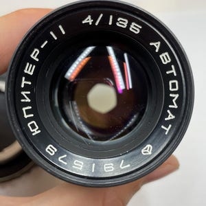 May include: A black camera lens with the text "4/135", "AUTOMAT", and "ЮНTEP-II" printed on the side. The lens has a black metal body and a silver ring around the edge. The lens is labeled with the number "761529".