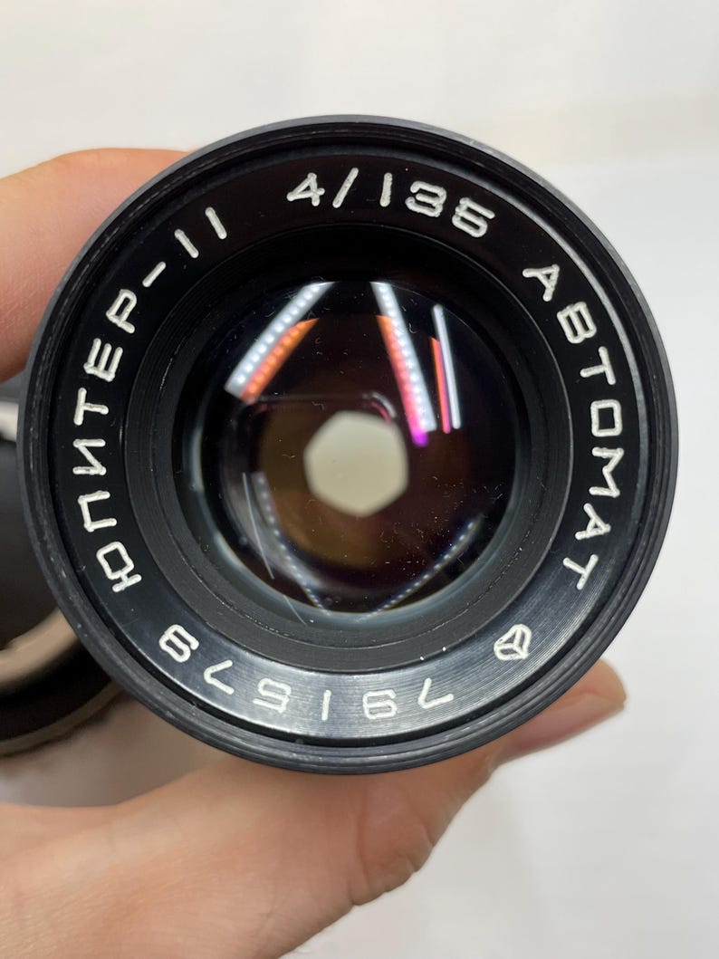 May include: A black camera lens with the text "4/135", "AUTOMAT", and "ЮНTEP-II" printed on the side. The lens has a black metal body and a silver ring around the edge. The lens is labeled with the number "761529".
