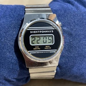以下が含まれることがあります： 金属製のリンクバンドと黒い長方形の文字盤を備えたシルバーカラーのデジタル腕時計。ディスプレイには22:05の時刻が表示されています。文字盤には「ЭЛЕКТРОНИКА 5」、「ЧАС」、「МИН」、「МЕСЯЦ」、「ЧИСЛО」、「СДЕЛАНО В СССР」という白い文字が記されています。