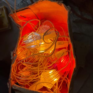 May include: A string of clear, faceted, diamond-shaped light bulbs emitting a warm orange glow, tangled inside a brown paper bag. The light illuminates the interior of the bag, highlighting the textured plastic surfaces of the bulbs and the thin, light-coloured electrical wiring connecting them together in a cluster.