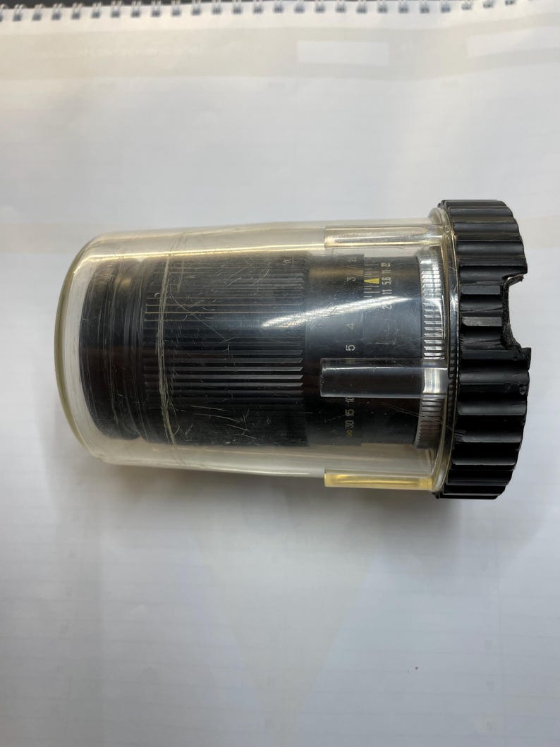 May include: A black camera lens with a clear plastic protective case. The lens has a silver ring with markings that include "50 1.5 30 5 1.5 30 5".