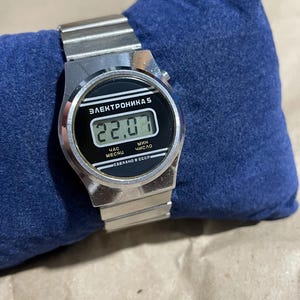 以下が含まれることがあります： 金属製のリンクバンドと黒い長方形の文字盤を備えたシルバーカラーのデジタル腕時計。ディスプレイには「22.07」という数字が表示されています。文字盤には「ЭЛЕКТРОНИКА 5」、「ЧАС」、「МЕСЯЦ」、「МИН」、「ЧИСЛО」、「СДЕЛАНО В СССР」という白い文字が記されています。