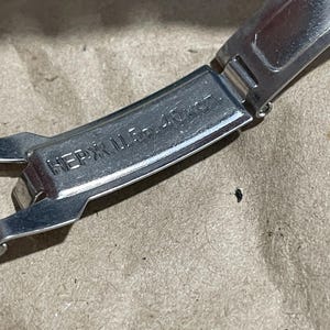 以下が含まれることがあります： 「НЕРЖ. Ц. 5р. 40коп.」と刻印されたシルバーカラーの金属製時計用クラスプ。クラスプは開いた状態で表示されており、金属の質感と内側に刻まれた文字がはっきりと確認できます。