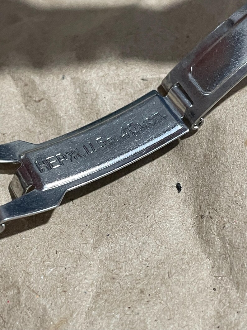 以下が含まれることがあります： 「НЕРЖ. Ц. 5р. 40коп.」と刻印されたシルバーカラーの金属製時計用クラスプ。クラスプは開いた状態で表示されており、金属の質感と内側に刻まれた文字がはっきりと確認できます。