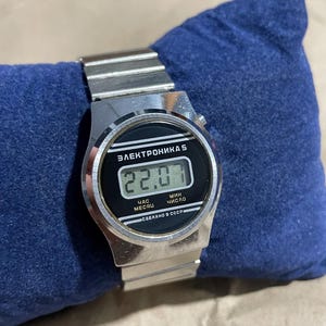 以下が含まれることがあります： 金属製のリンクバンドと黒い長方形の文字盤を備えたシルバーカラーのデジタル腕時計。ディスプレイの上部には「ЭЛЕКТРОНИКА 5」、数字の下には「ЧАС МЕСЯЦ」と「МИН ЧИСЛО」、下端には「СДЕЛАНО В СССР」という文字が表示されています。