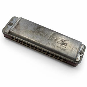 May include: A vintage harmonica with a silver metal casing. The word "Perfecta" is engraved on the top, along with the phrase "Fur Kenner". The harmonica has a rectangular shape with multiple holes and a red accent.