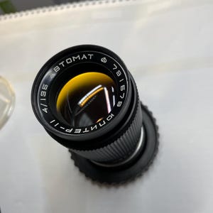 Könnte beinhalten: Ein schwarzes Kameraobjektiv mit dem Text "AUTOMAT" und "ЮНТЕП-II" darauf gedruckt. Das Objektiv hat einen schwarzen Metallkörper und einen silbernen Ring um die Vorderseite.