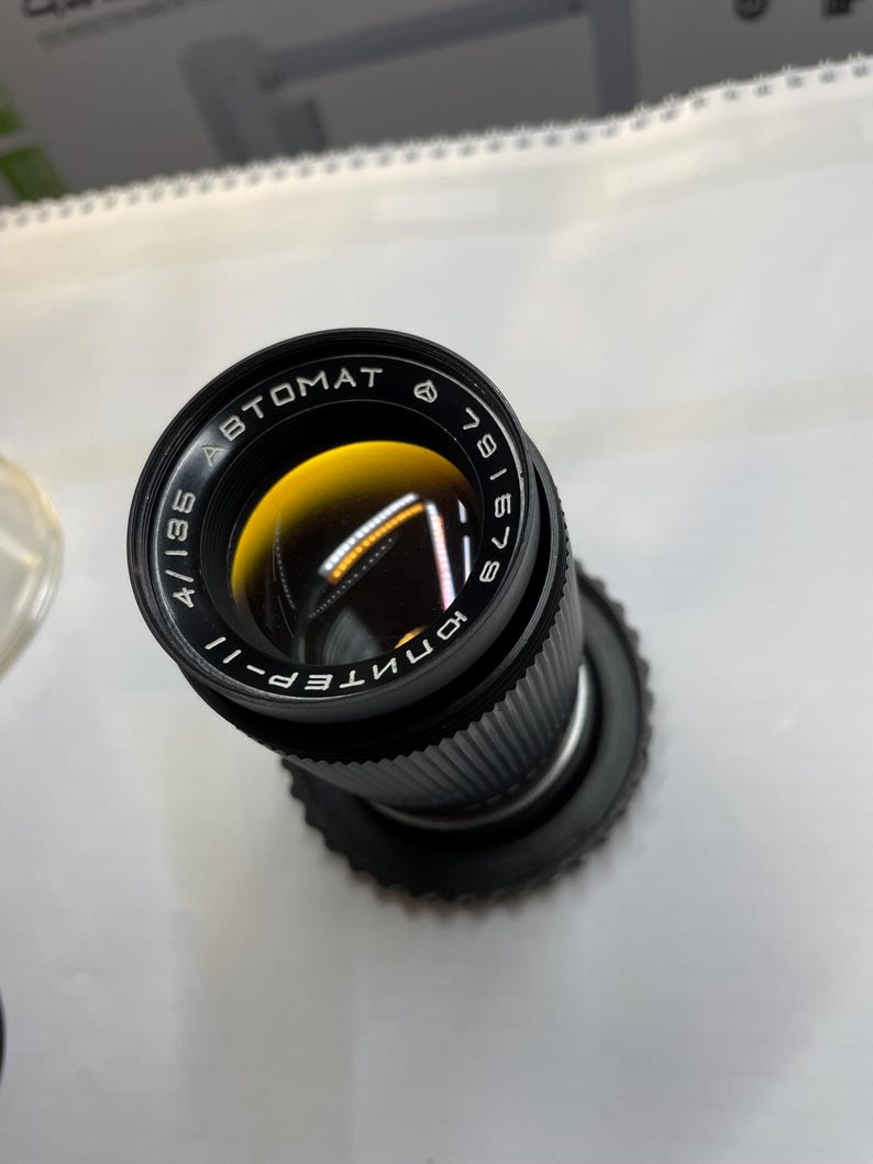 May include: A black camera lens with the text "AUTOMAT" and "ЮНТЕП-II" printed on it. The lens has a black metal body and a silver ring around the front.