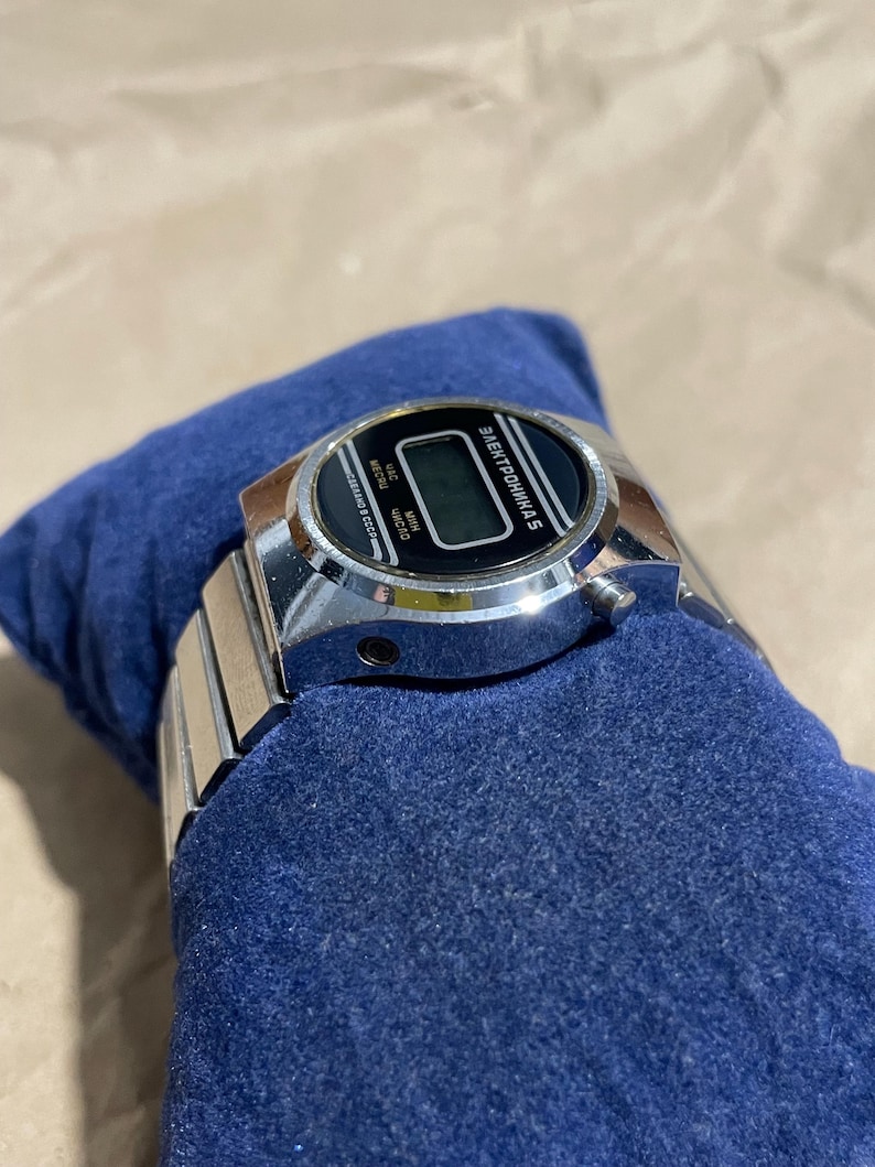 以下が含まれることがあります： 金属製のリンクバンドと黒い長方形のディスプレイ面を備えたシルバーカラーのデジタル腕時計。時計の文字盤にはキリル文字で「ЭЛЕКТРОНИКА 5」および「СДЕЛАНО В СССР」と印字されており、デジタル時刻表示インジケーターも備わっています。