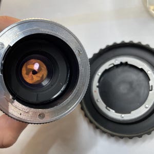 May include: Close-up of a silver camera lens with a black ring around the lens. The lens is showing the inside of the lens with a circular opening. The lens is sitting on a white surface.