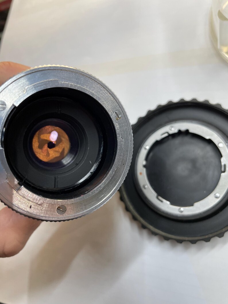 May include: Close-up of a silver camera lens with a black ring around the lens. The lens is showing the inside of the lens with a circular opening. The lens is sitting on a white surface.