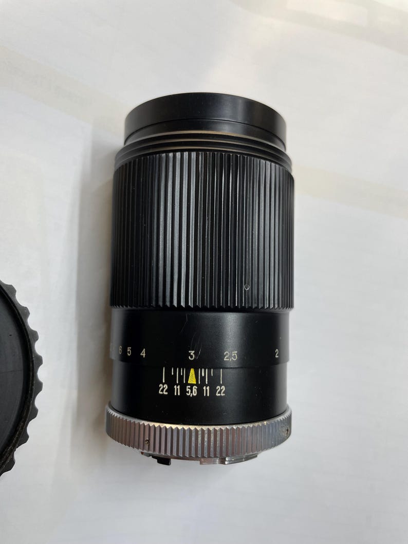 May include: A black camera lens with a silver ring and a yellow triangle marking the aperture setting. The lens has a depth of field scale marked with numbers from 2 to 6 and aperture settings from 22 to 5.6.