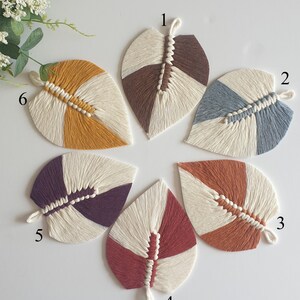 May include: Six handmade macrame leaf wall hangings in various colours, including brown, blue, yellow, purple, red, and orange. Each leaf is made with white cotton cord and features a unique colour combination.