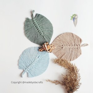 May include: Three macrame leaf wall hangings in blue, green, and beige colours. The leaves are arranged in a triangular shape with a seashell in the centre.